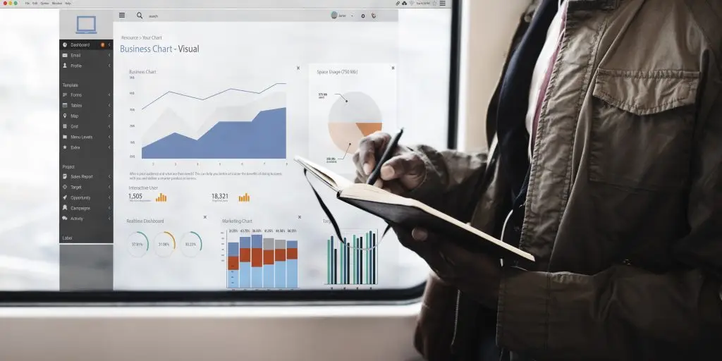 Persona tomando notas en una libreta mientras analiza gráficos y estadísticas de un panel de control empresarial en un entorno digital, representando análisis de datos y toma de decisiones